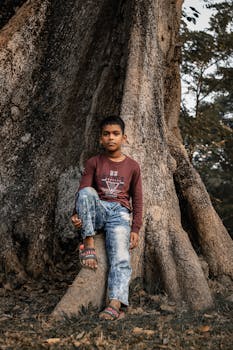 A young boy in casual attire leans against the massive roots of an ancient tree in a rural setting.