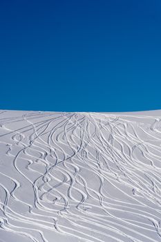 Snowy ski slopes with intricate ski tracks against a clear blue sky on a bright winter day.