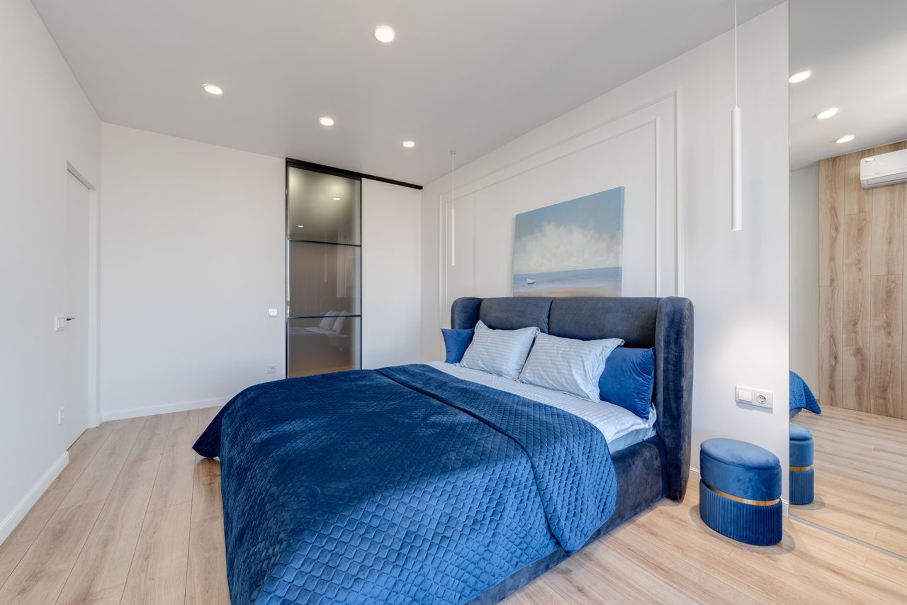 Wide view of a bedroom with a deep navy blue velvet duvet, white pillows and light wood flooring