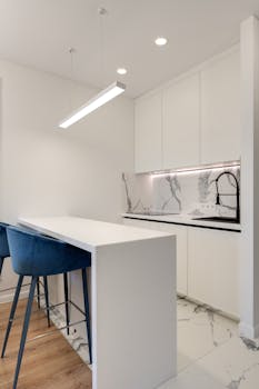 Sleek white kitchen with marble counter and blue bar stools, showcasing modern design.
