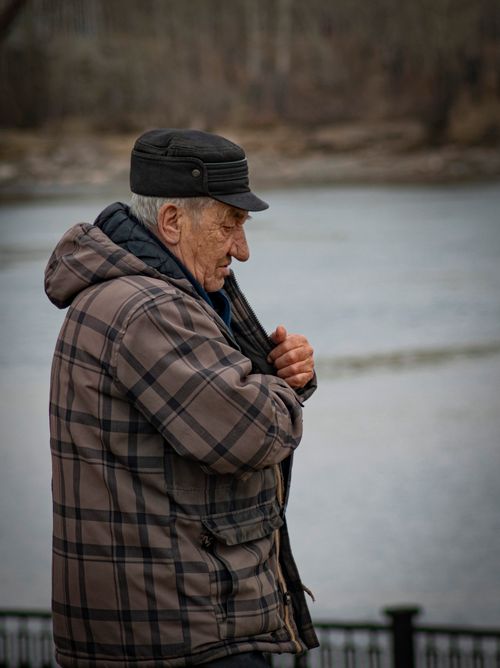Free An elderly man in a plaid jacket standing pensively by a river with a blurred background. Stock Photo