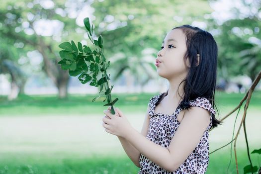 A young girl with dark hair holds a leafy branch outdoors in a sunlit park.