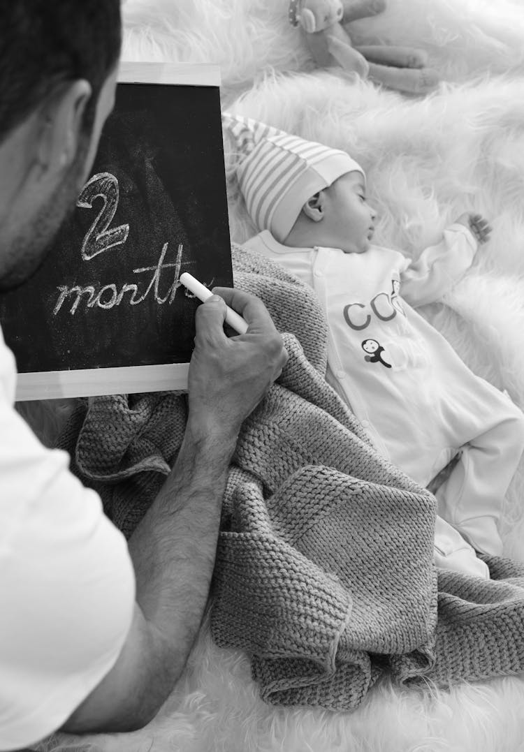 A Man Writing On The Blackboard Near His Baby Sleeping On A Fur Carpet