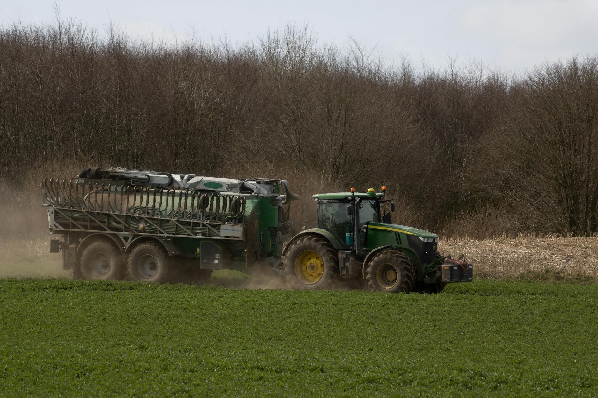 A green tractor pulling a trailer across a green field against a backdrop of trees in rural landscape.