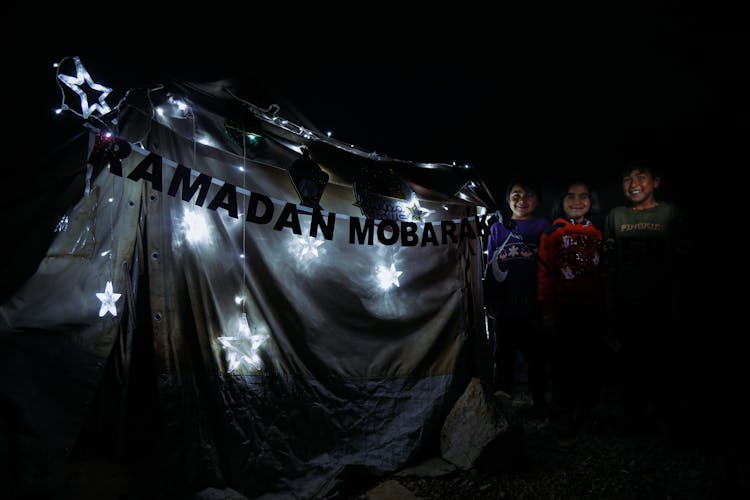 A Group Of Kids Standing Beside Ramadan Mobarak Signage With String Lights