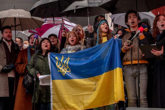 A diverse group under umbrellas holding a Ukrainian flag and singing outdoors.