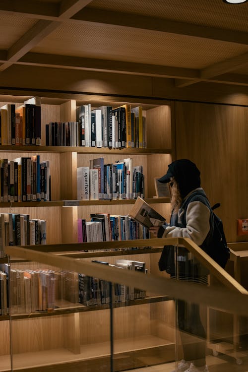 A Person Reading a Book Near the Wooden Shelves