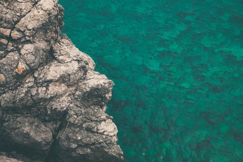 Free Aerial view of rugged coastal rocks meeting the clear turquoise ocean water on a sunny day. Stock Photo