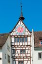 Old dark brown medieval clock tower with cute weather vane on a white, half timbered tenement house with pastel colored and golden round clock on sunny day