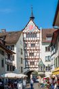 Old dark brown medieval clock tower with cute weather vane on a white, half timbered tenement house with pastel colored and golden round clock on sunny day
