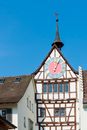 Old dark brown medieval clock tower with cute weather vane on a white, half timbered tenement house with pastel colored and golden round clock on sunny day