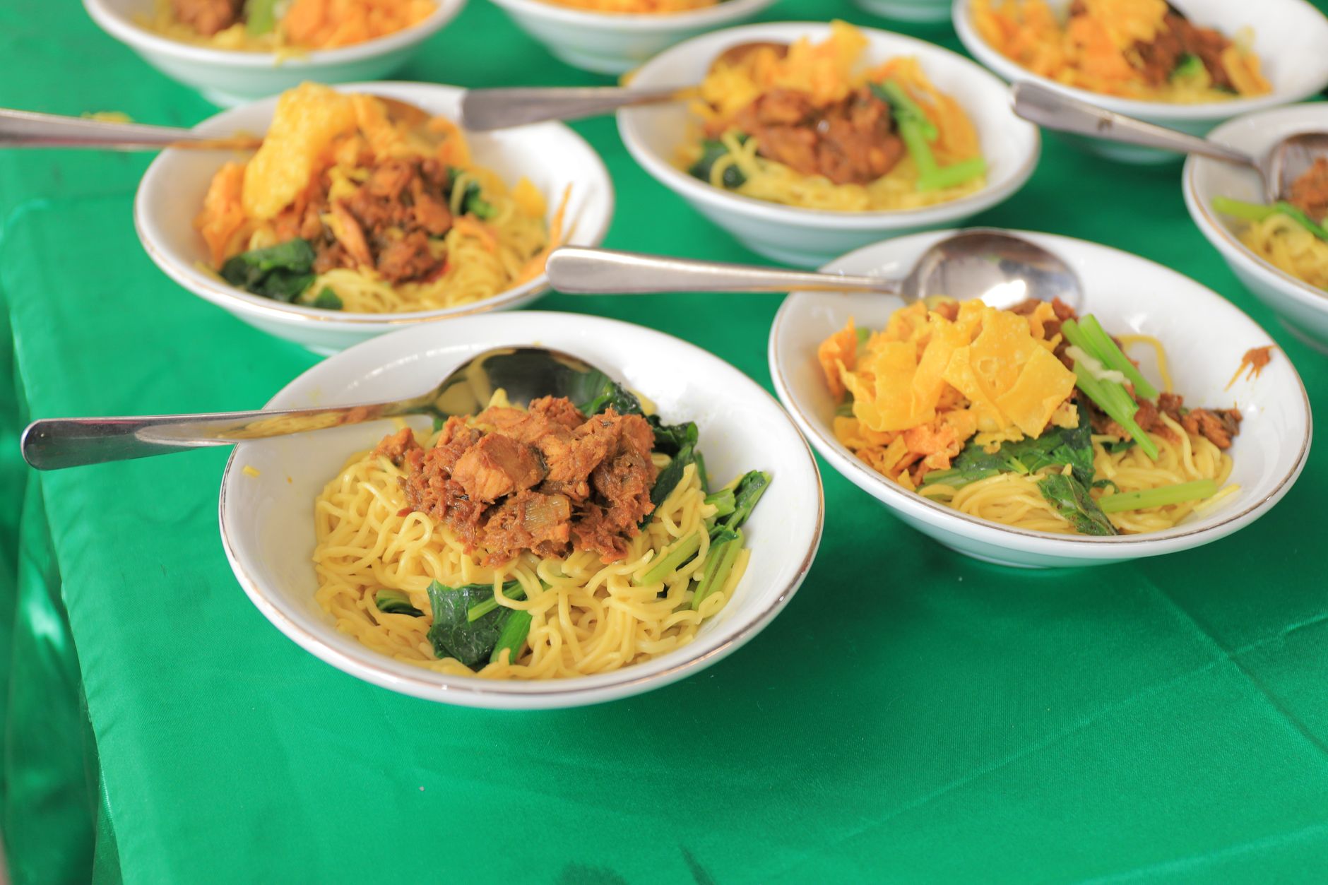 Beragam mangkuk mie Asia dengan daging dan sayuran di atas taplak meja hijau, disajikan dengan sendok.