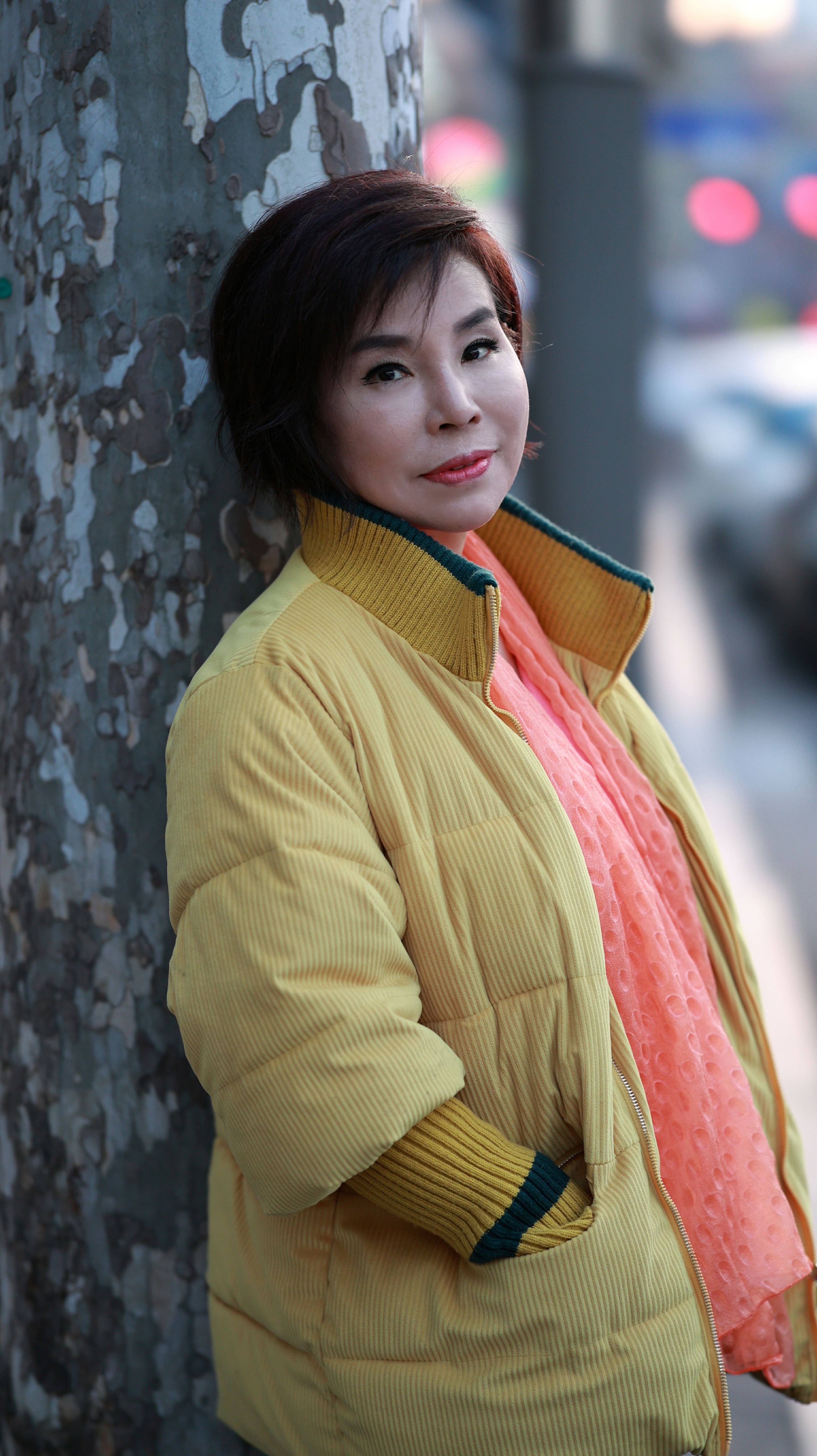 Asian woman in a yellow jacket outside, leaning on a tree trunk, looking at camera.
