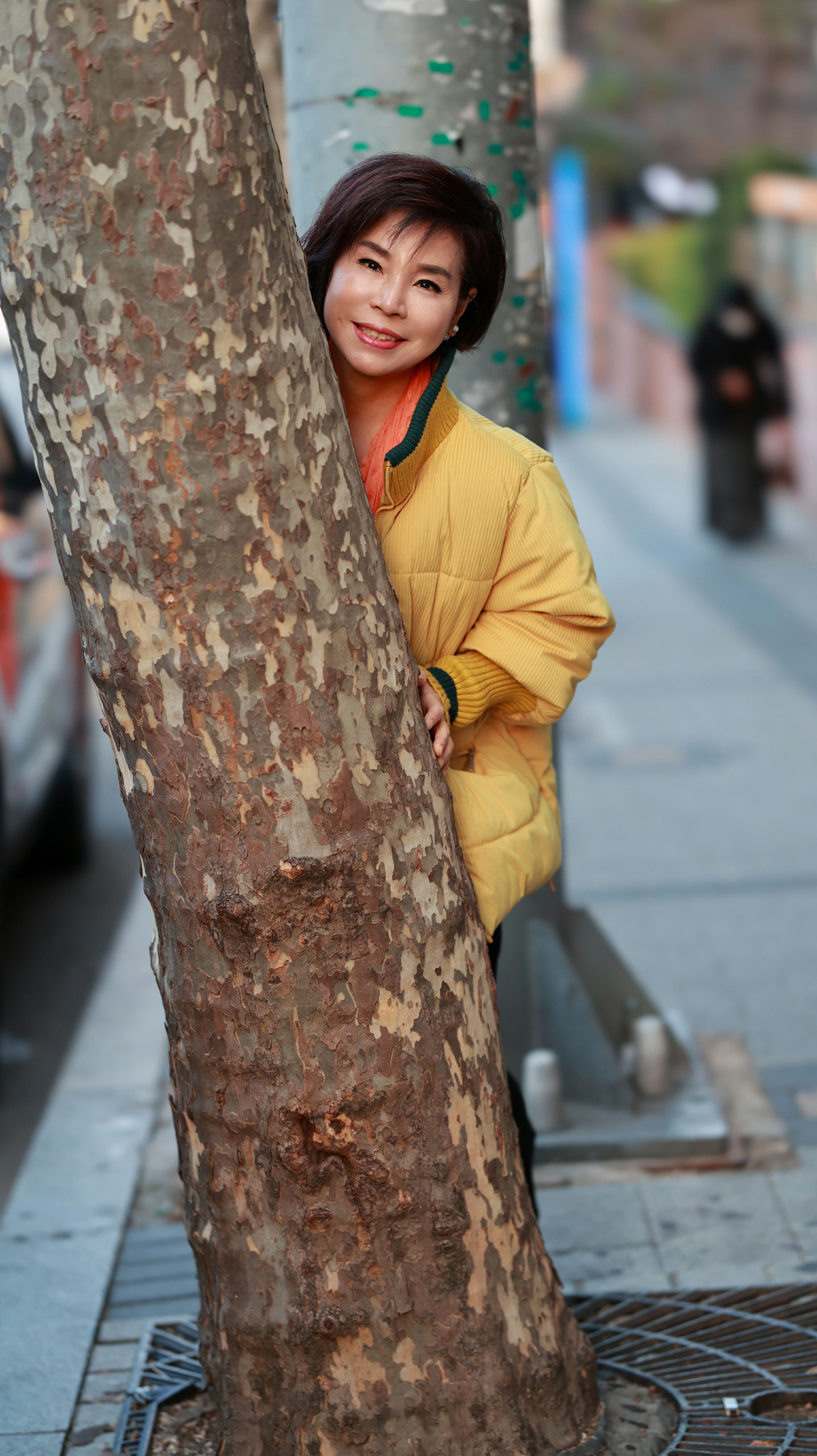 A cheerful Asian woman in a yellow jacket leans playfully against a tree in an urban setting.