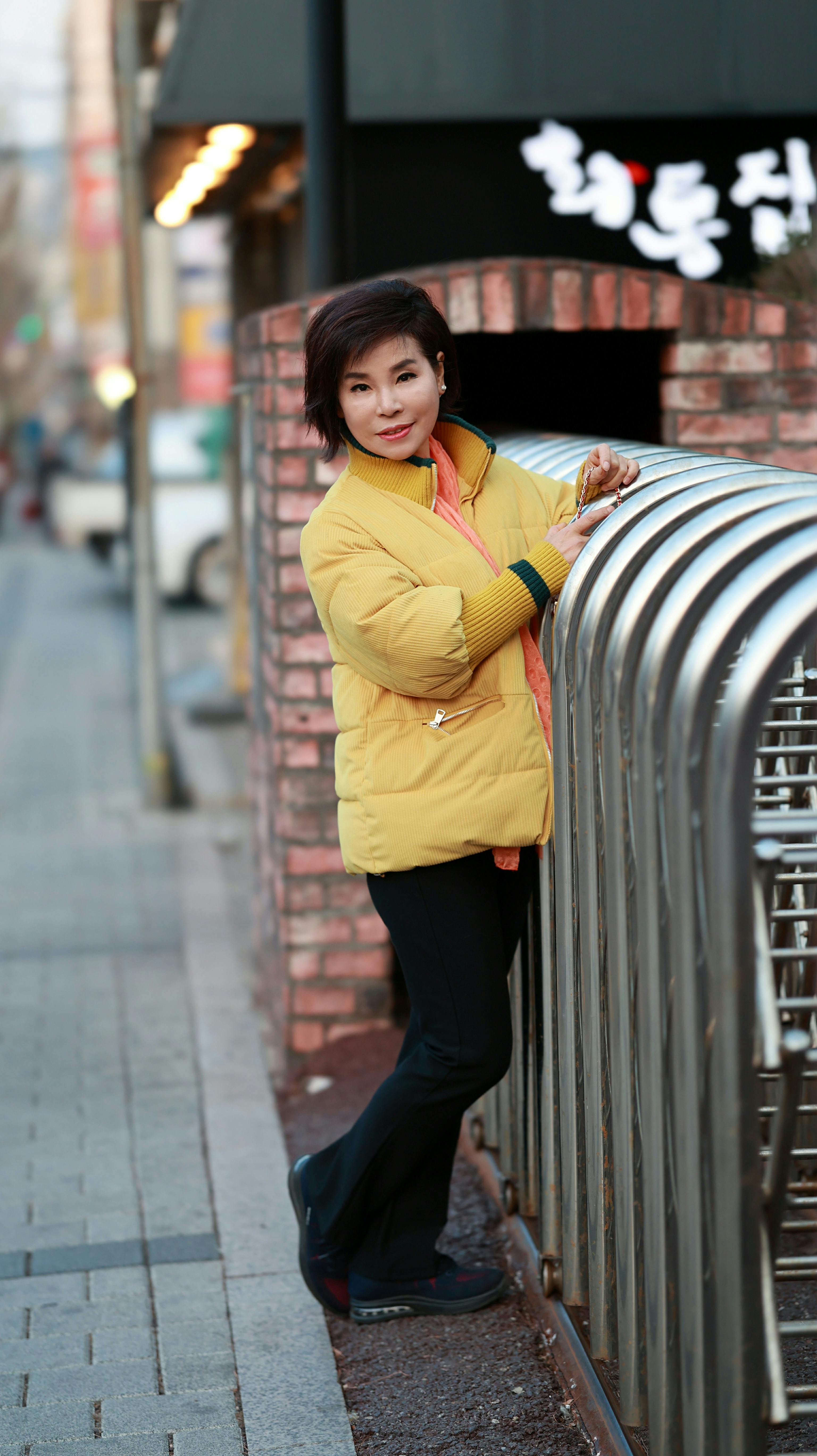 Smiling Asian woman standing by metal railings on a city street wearing a yellow jacket.