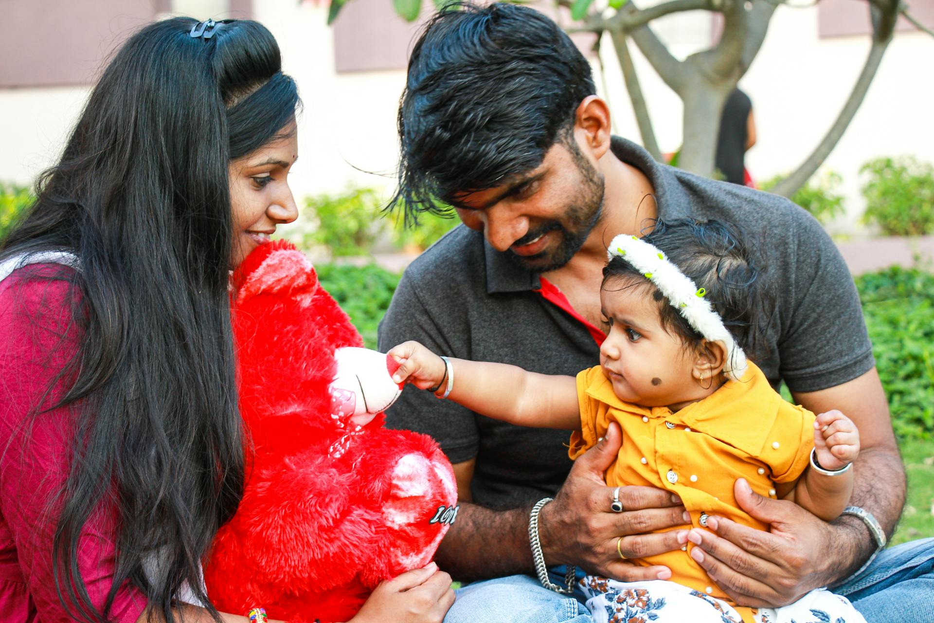 Indian parents and their toddler enjoy outdoor time with a bright red teddy bear.