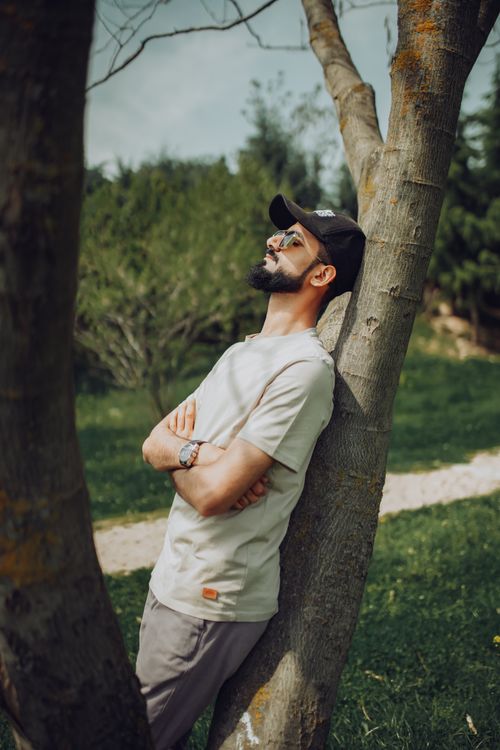 Free A bearded man in a beige shirt leans against a tree outdoors in a sunlit park, embracing tranquility. Stock Photo