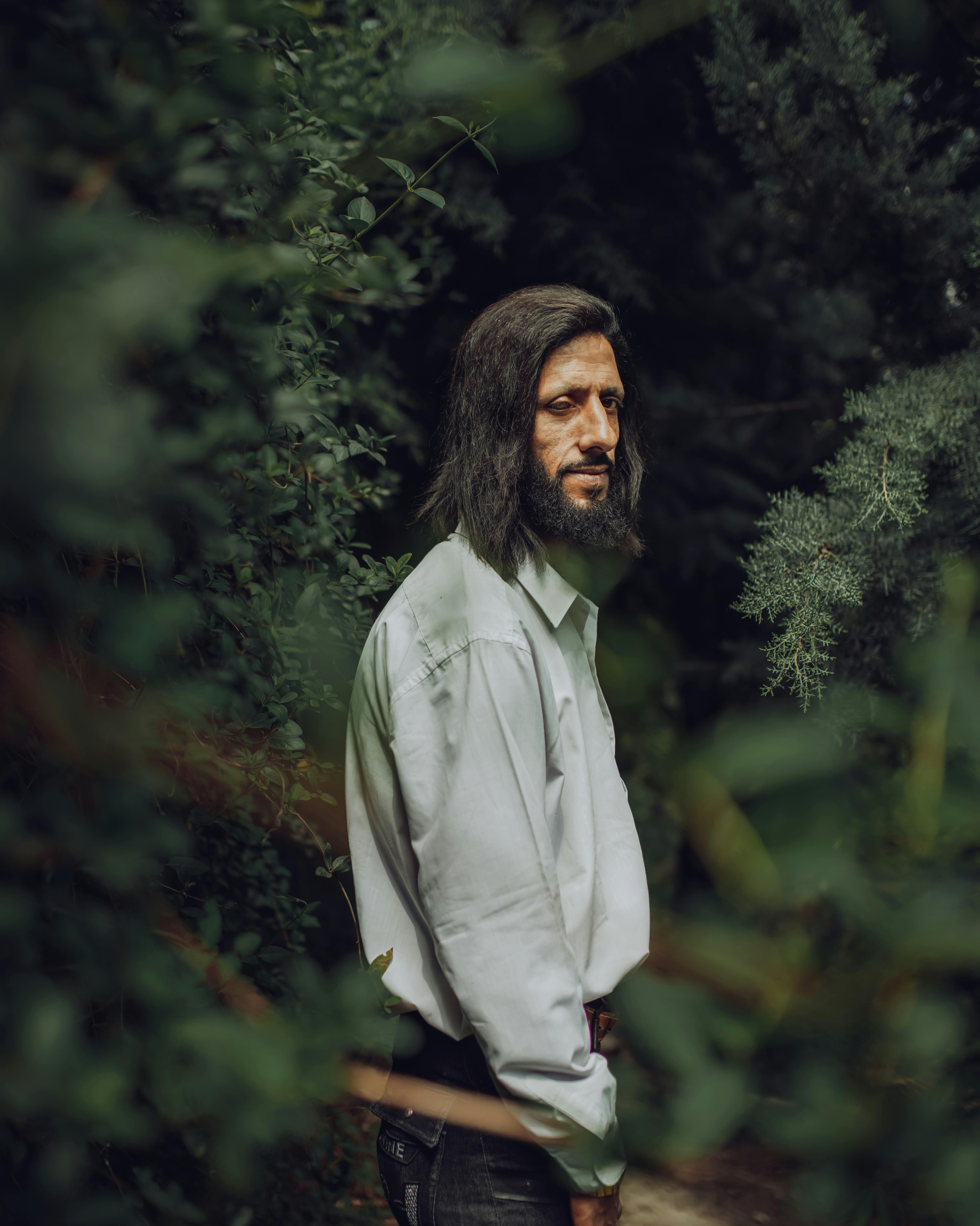 Man with long hair and beard standing in green foliage, wearing a white shirt.