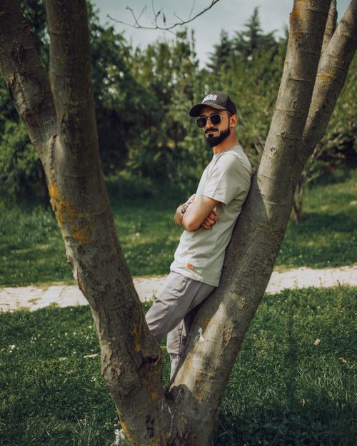 Free A man in a cap and sunglasses leans on a tree in a lush green park. Stock Photo