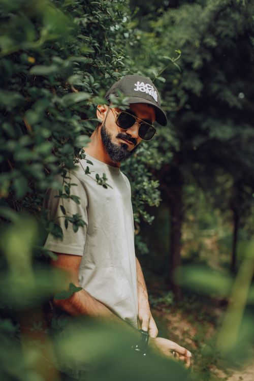 Free Stylish bearded man with sunglasses and cap posing among greenery during the day. Stock Photo