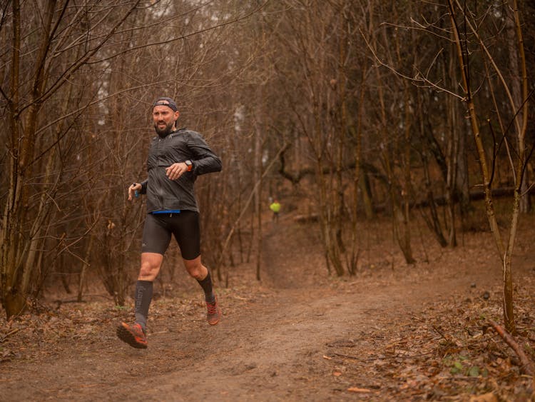 A Man In Black Jacket And Cycling Shorts Running Near The Leafless Trees