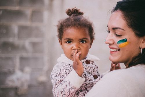 Free A smiling woman with face paint holds a toddler with a thoughtful look outdoors. Stock Photo
