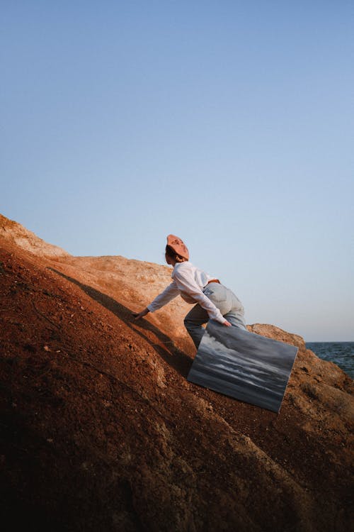 Unrecognizable Woman Climbing Red Rock with Painting in Hand