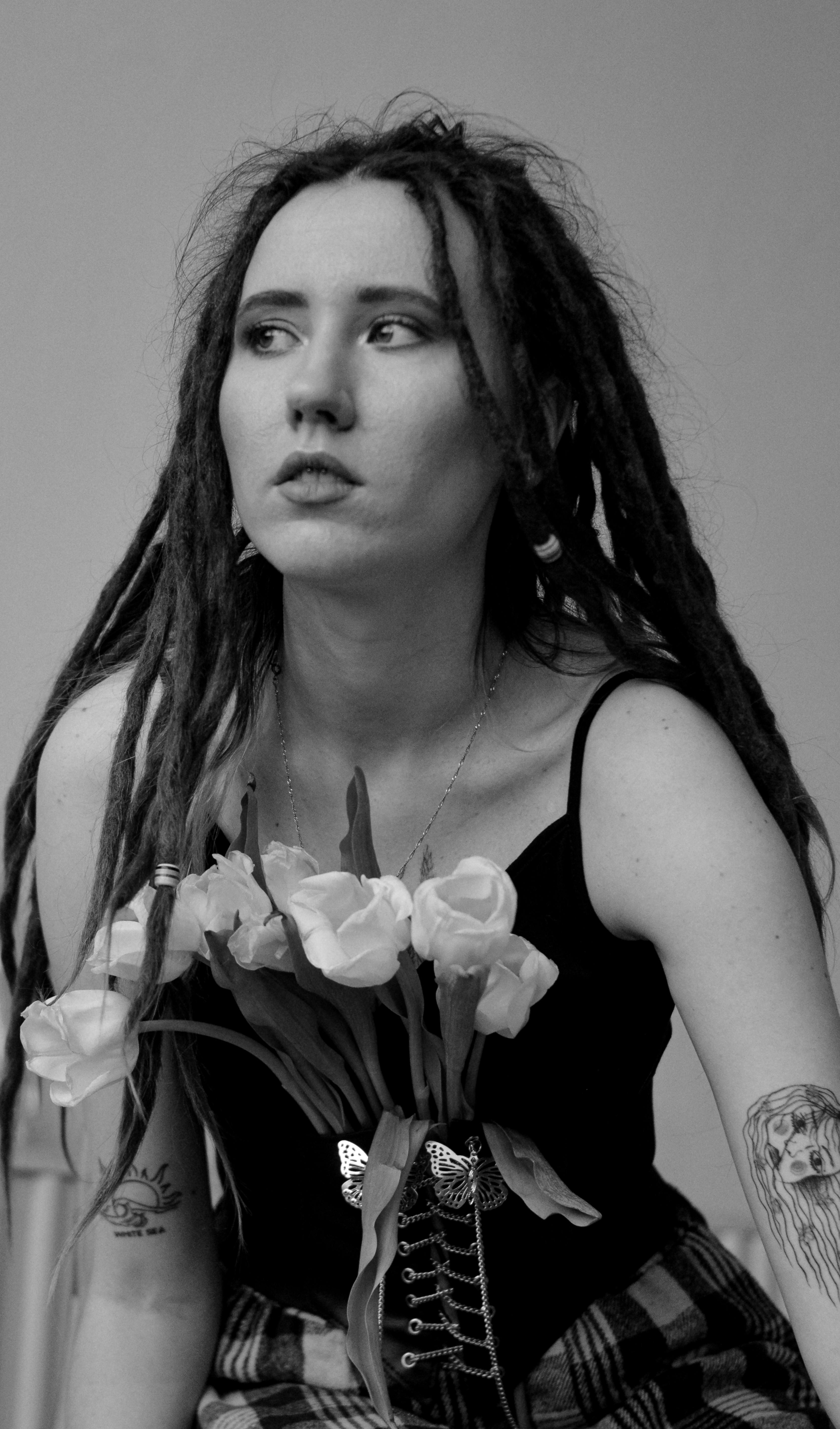 Artistic black and white portrait of a woman with dreadlocks, tattoos, and a corset holding flowers.