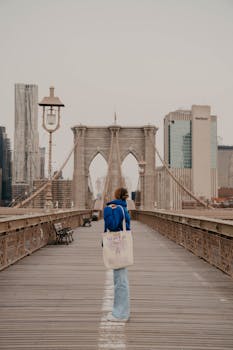 A woman in casual attire walks on Brooklyn Bridge, surrounded by New York City's skyline.