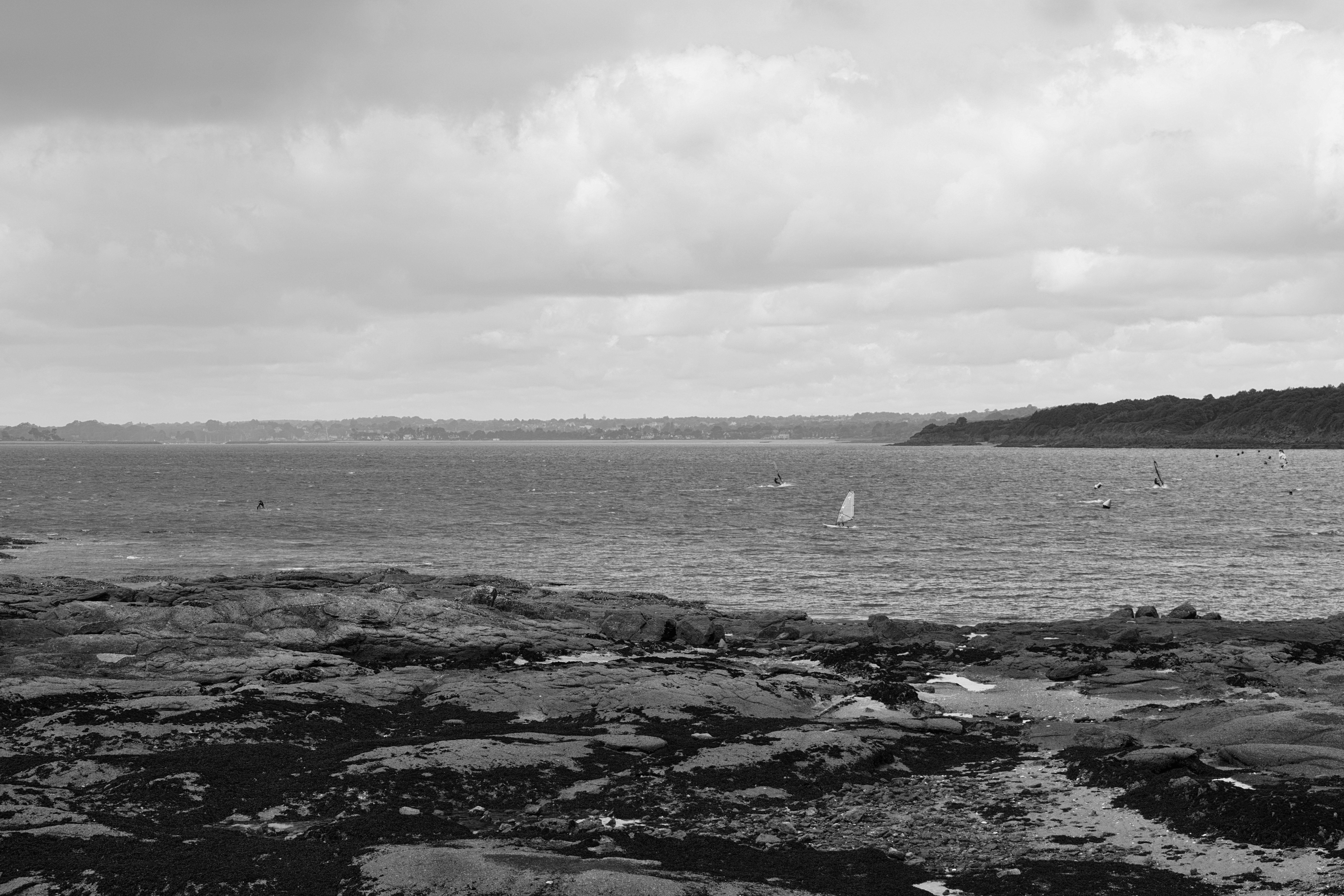 Calm sea with rocky shoreline under cloudy skies, featuring distant windsurfers.