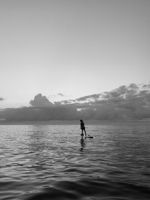 Free A lone paddleboarder glides on calm ocean waters under a dramatic sky at sunset. Stock Photo