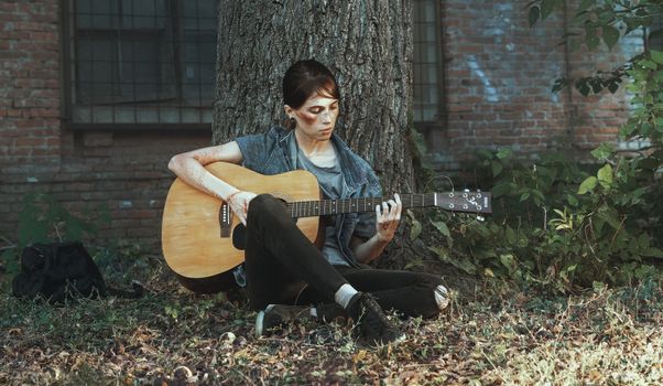 A woman sitting against a tree, playing an acoustic guitar in a peaceful outdoor setting.