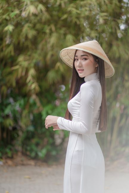 Elegantly dressed woman in a white ao dai and conical hat, smiling outdoors.