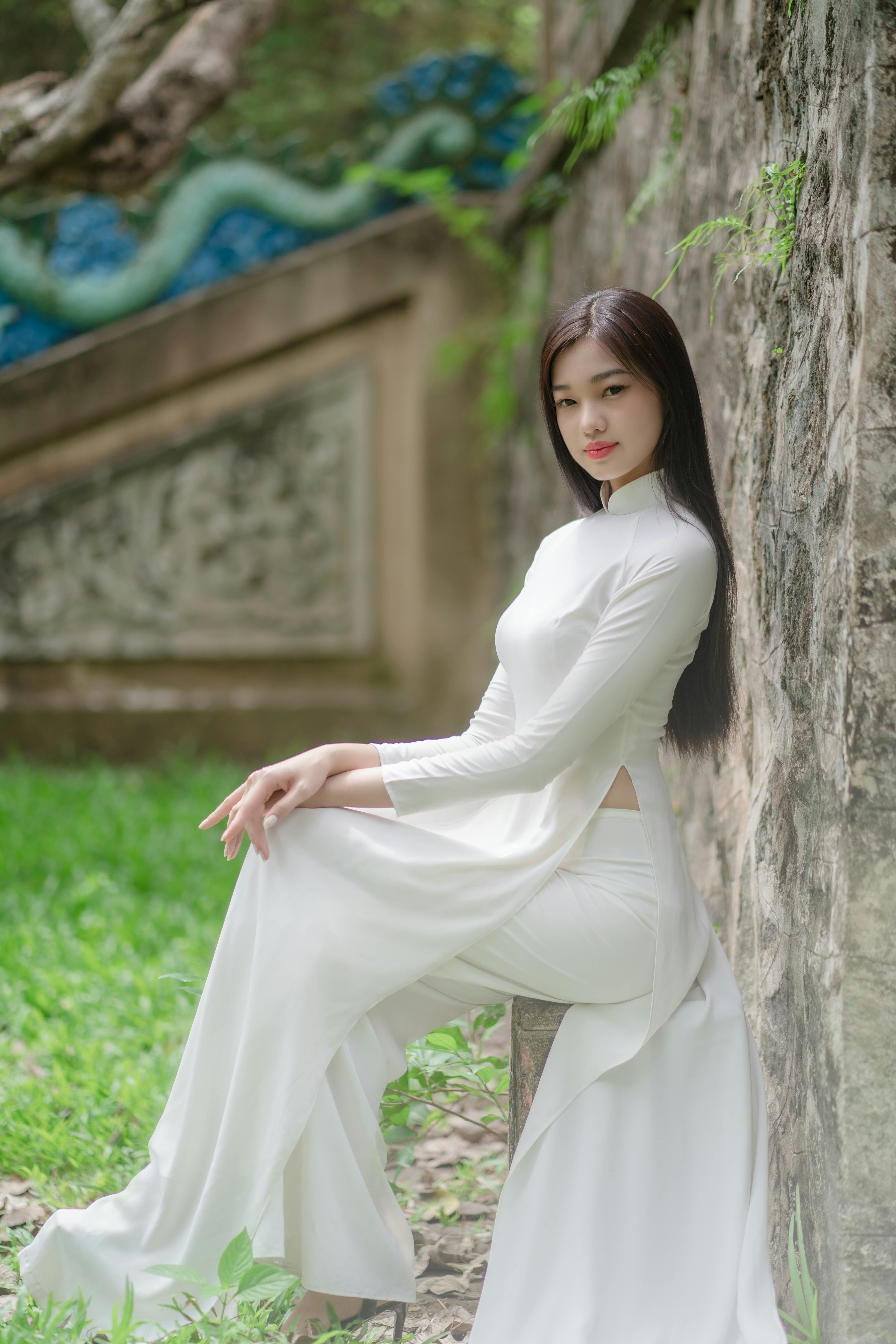 A woman in a traditional white Ao Dai gracefully sitting outdoors by a stone wall.