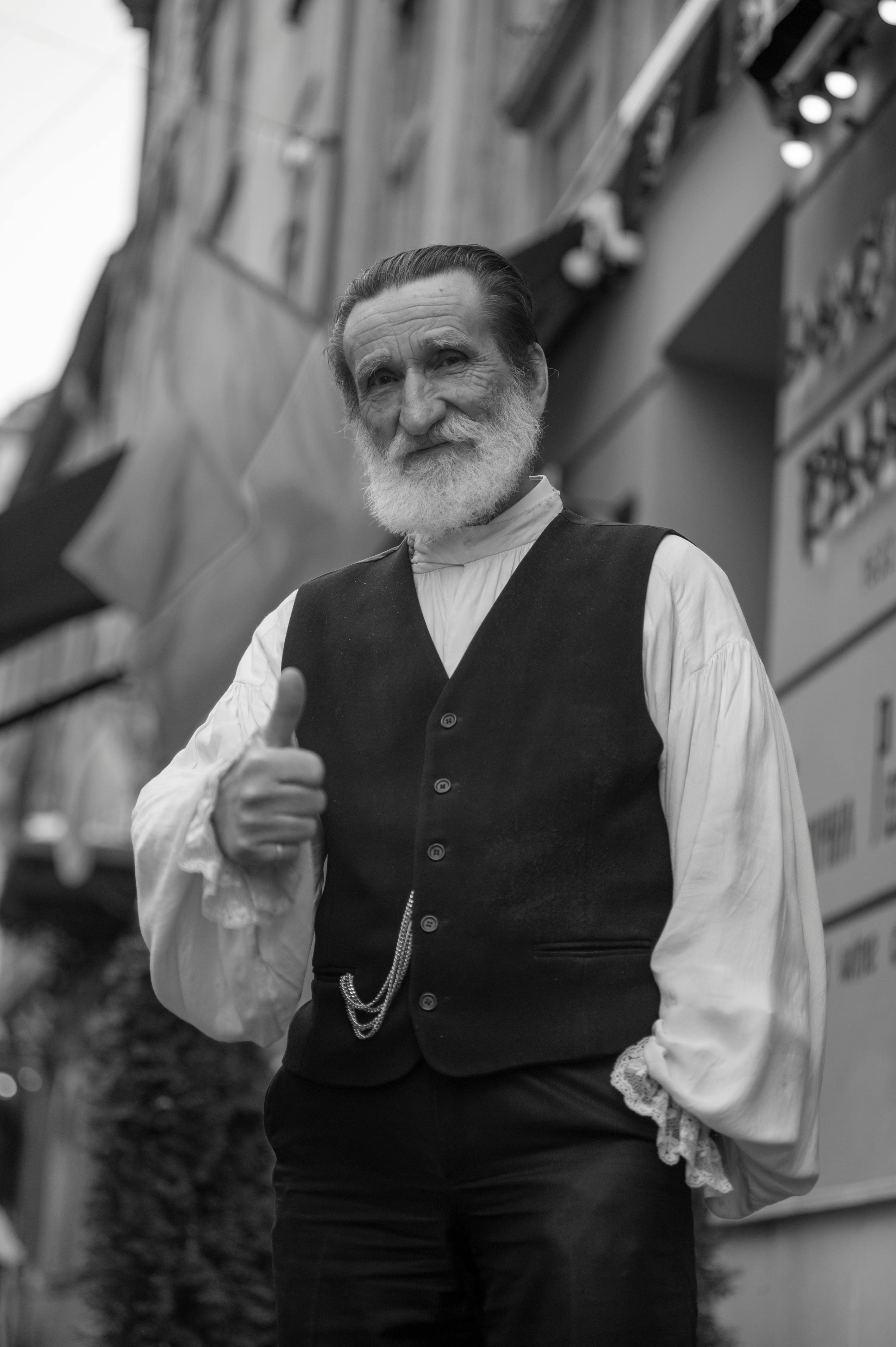 A senior man with a white beard gives a thumbs up in a street of Lviv, wearing traditional attire.