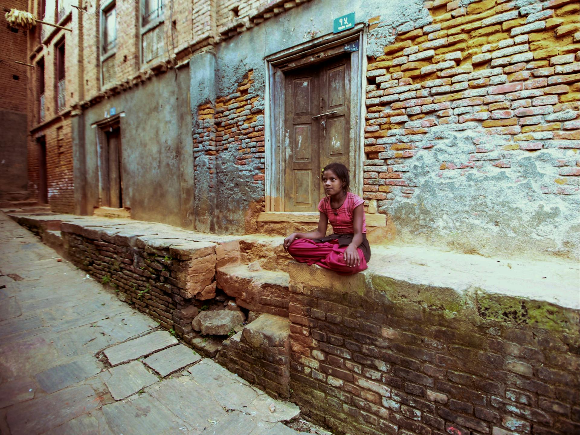 A young girl sits alone on a brick wall in a slum area, reflecting poverty and solitude.