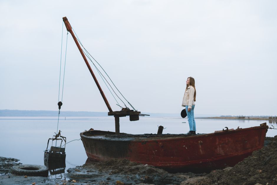 A woman stands on an old, rusty boat at a serene lakeside, capturing a moment of contemplation.