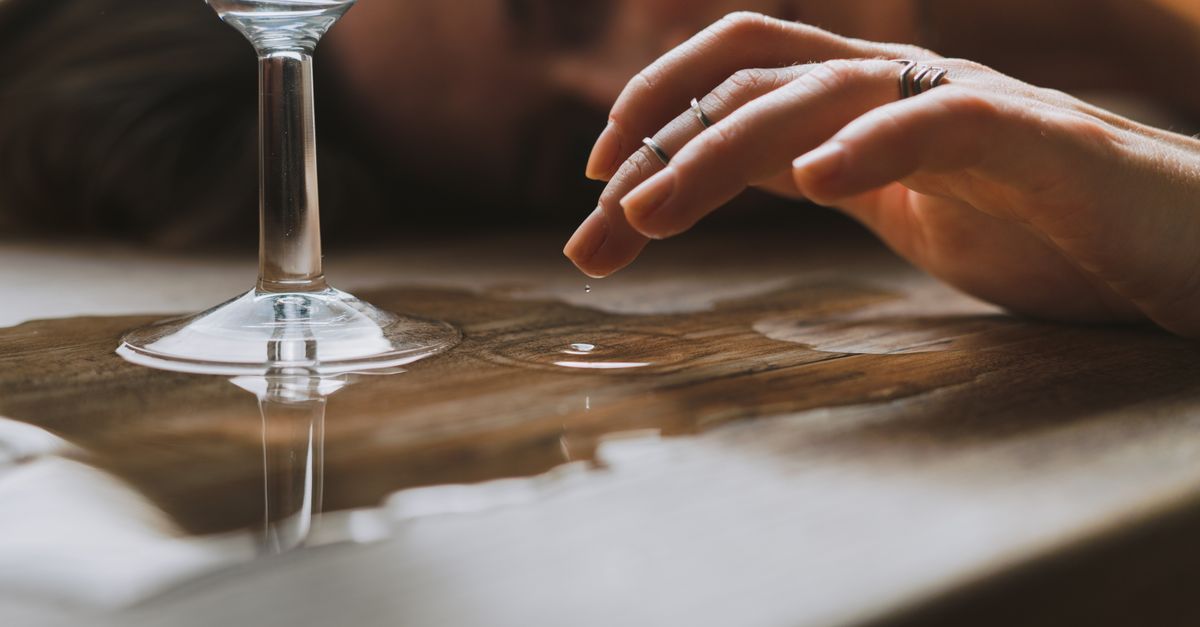 Uma imagem aproximada de uma mão tocando uma gota de água ao lado de um copo em uma superfície de madeira.
