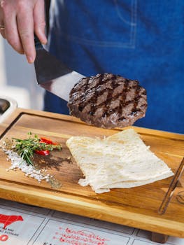 A grilled hamburger patty on a spatula with herbs, spices, and flatbread on a wooden tray.