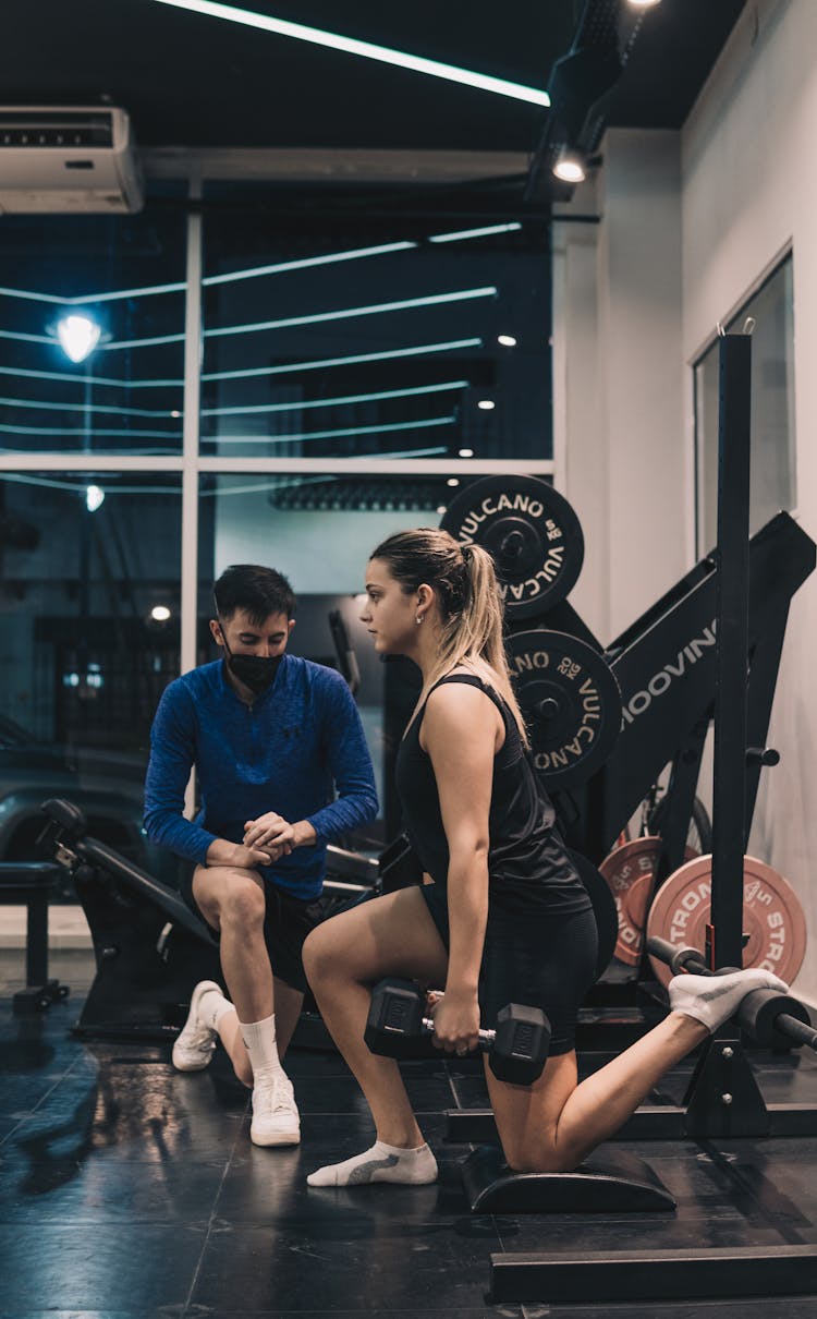 Young Woman Kneeling With Exercise Dumbbell In Hand And Man Accompanying Her