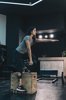 Focused woman exercises with dumbbells on a wooden box in a modern gym.