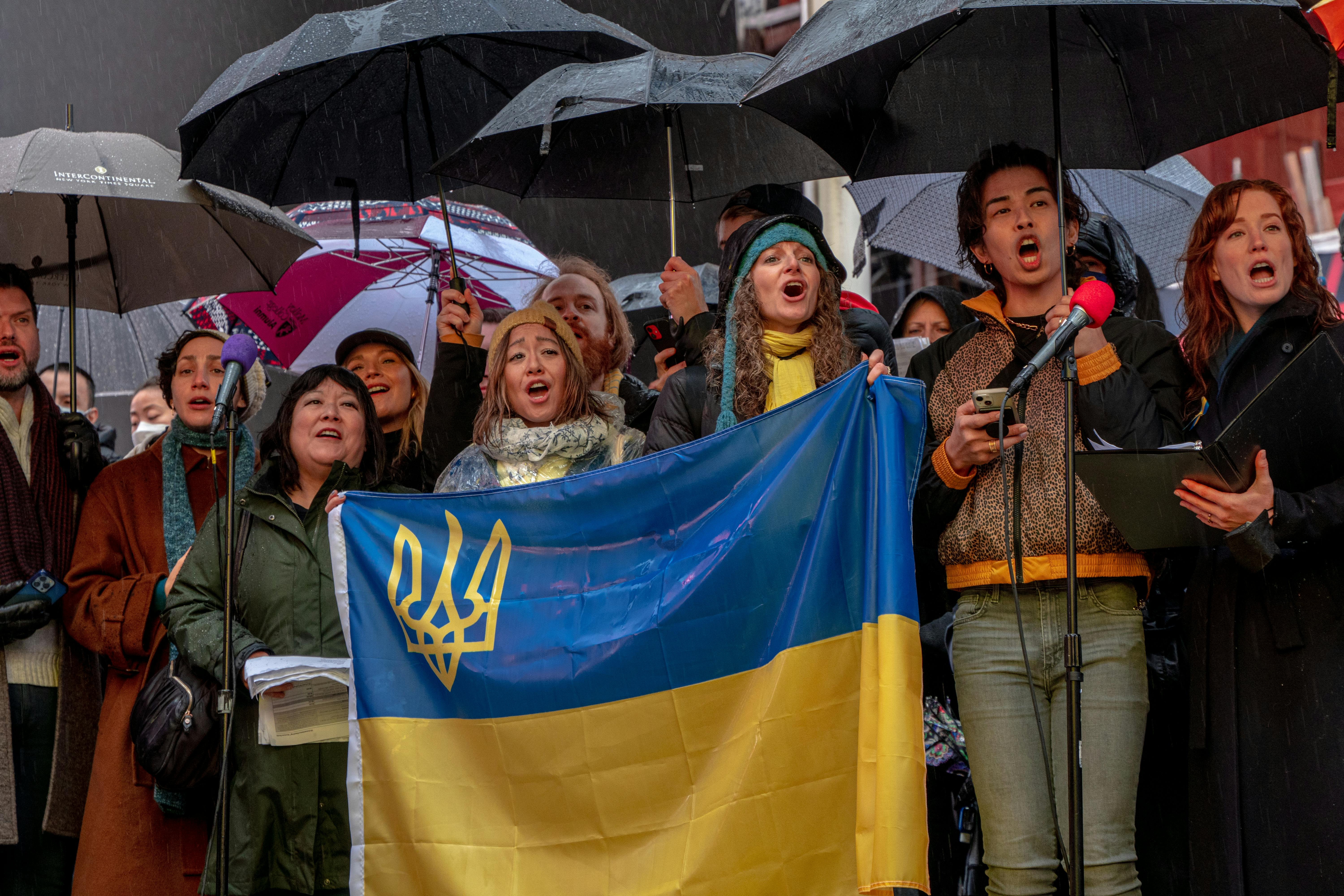 Diverse group at a support rally in New York, holding Ukrainian flag and umbrellas.