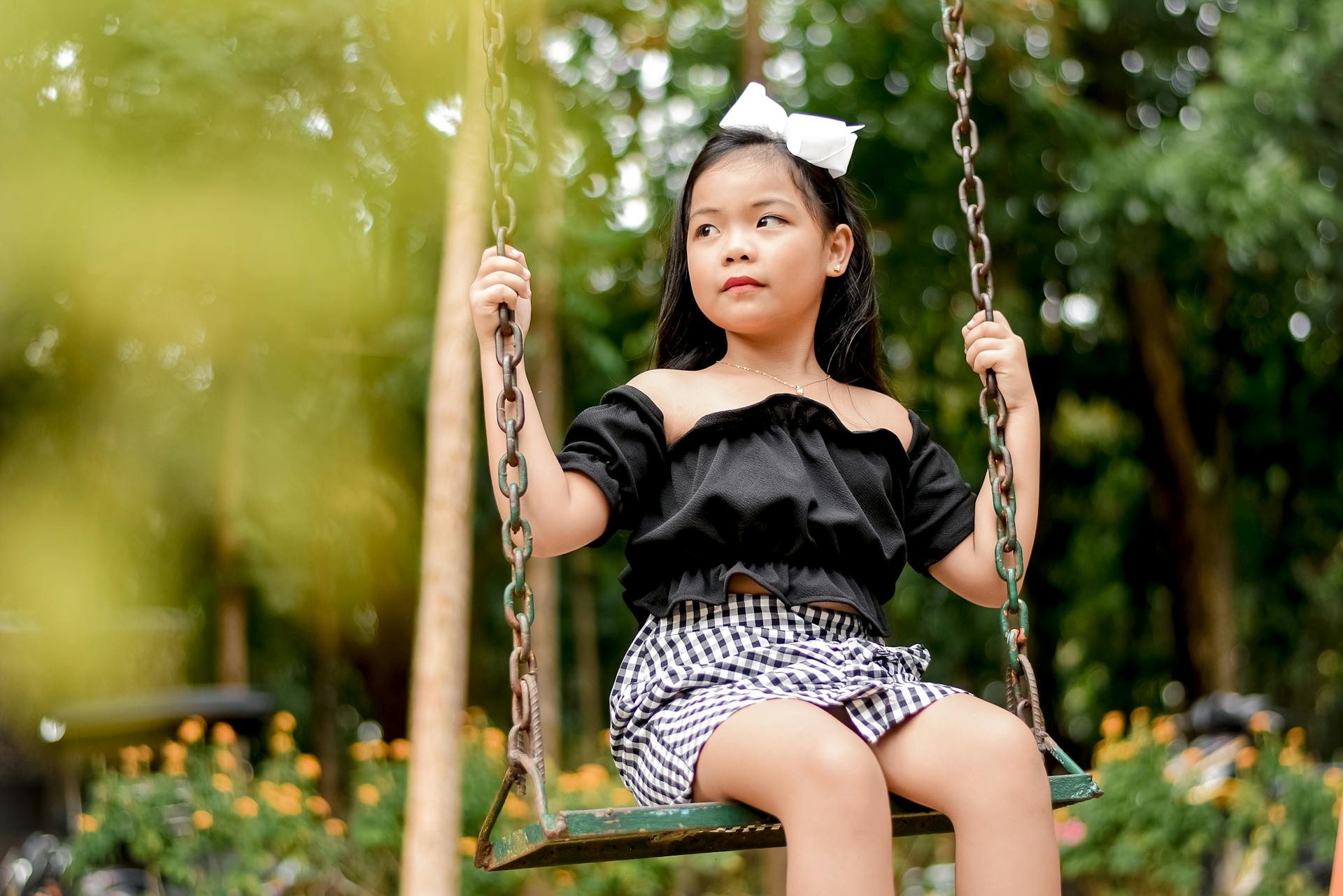 Cute little girl in fashionable attire sitting on a swing outdoors, looking away with a thoughtful expression.