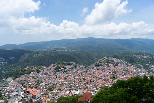 A panoramic view of Taxco, Mexico, with vibrant rooftops and lush mountains under a bright blue sky.