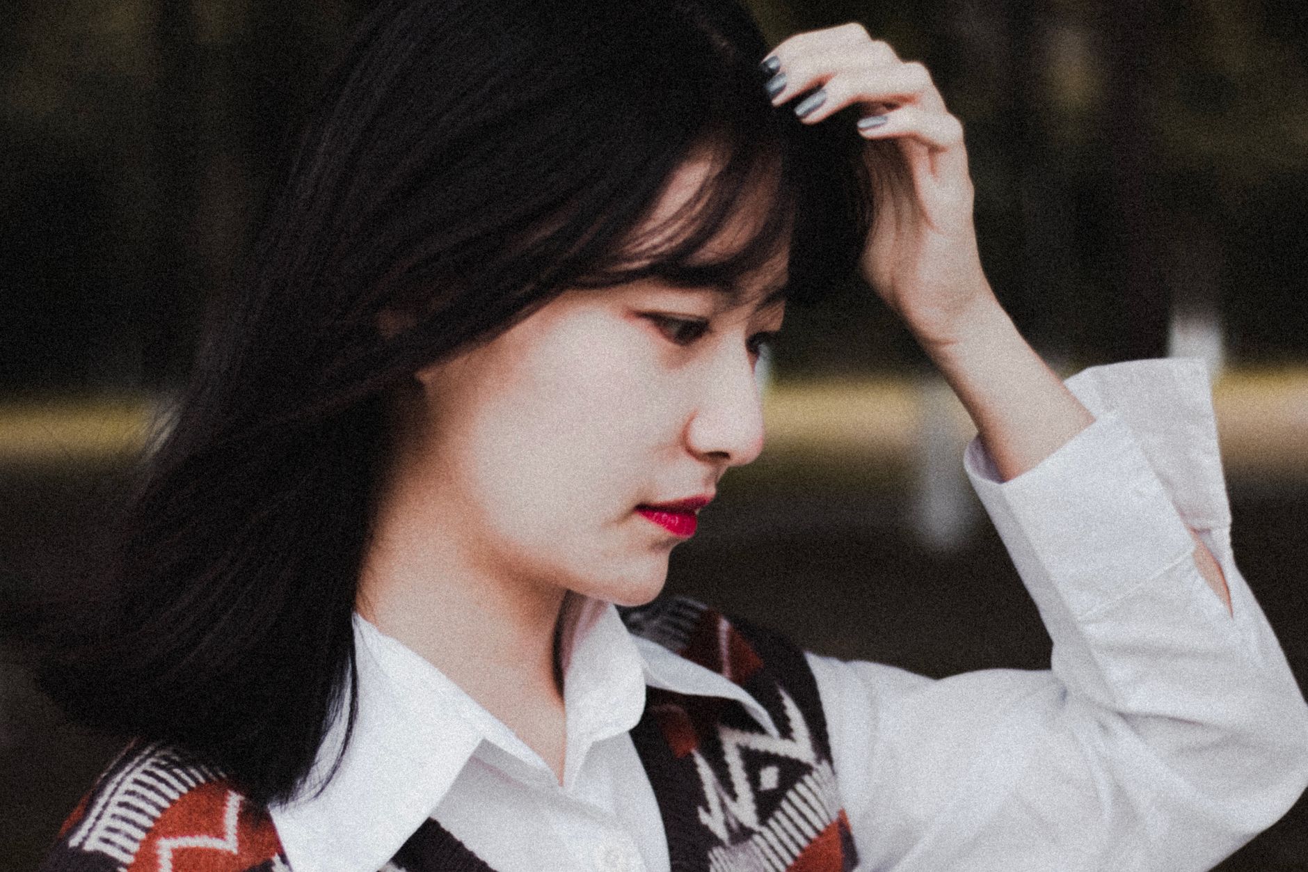 Elegant woman with pink lips and dark hair adjusting her hairstyle while outdoors, expressing style and grace.
