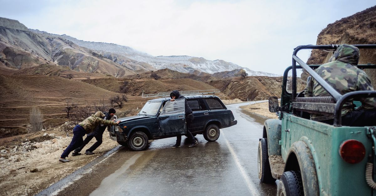 Men pushing a stuck SUV on a wet mountain road while another jeep waits nearby.