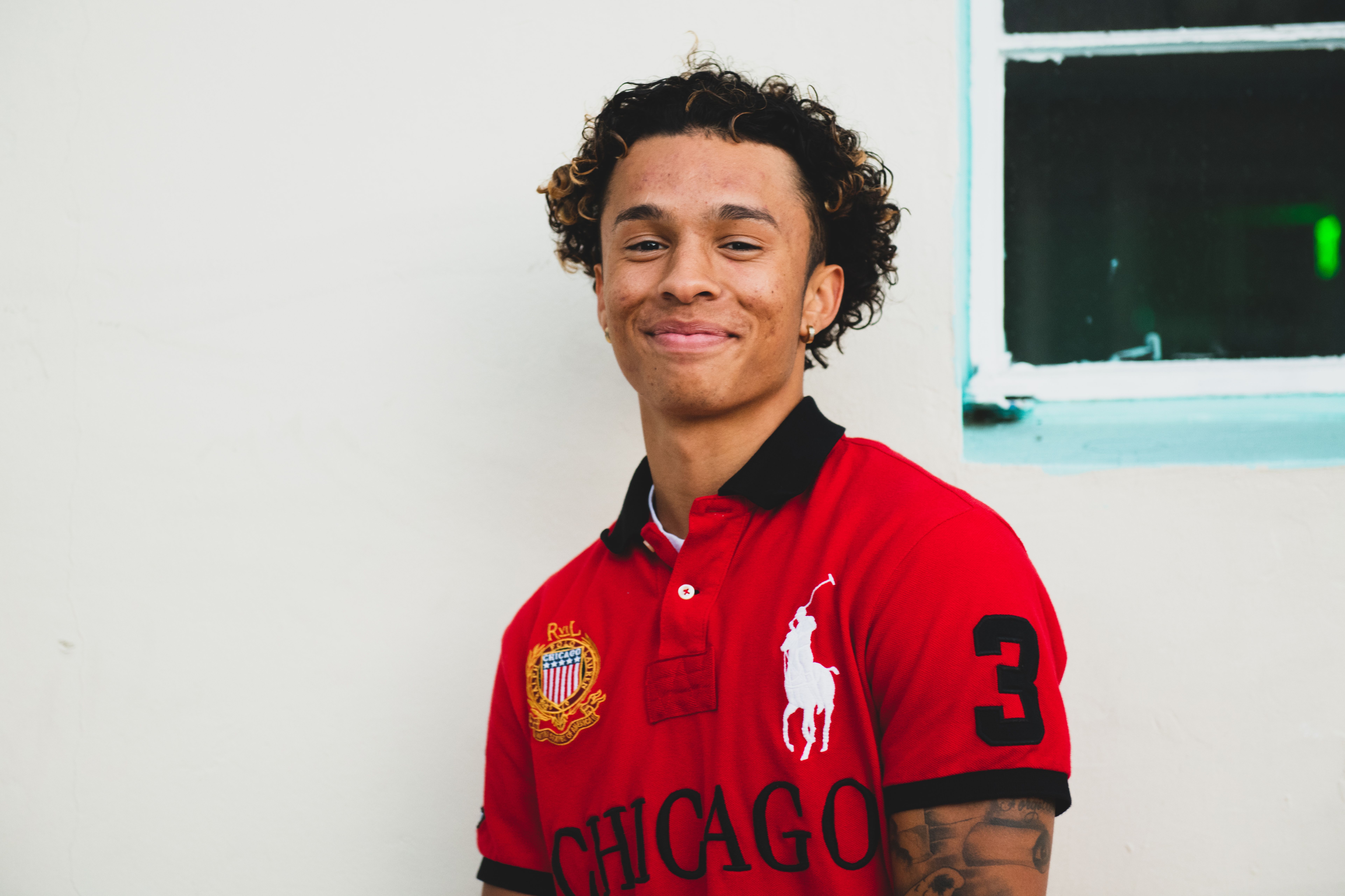 Young man smiling in a red polo shirt with tattoos, standing by a building.