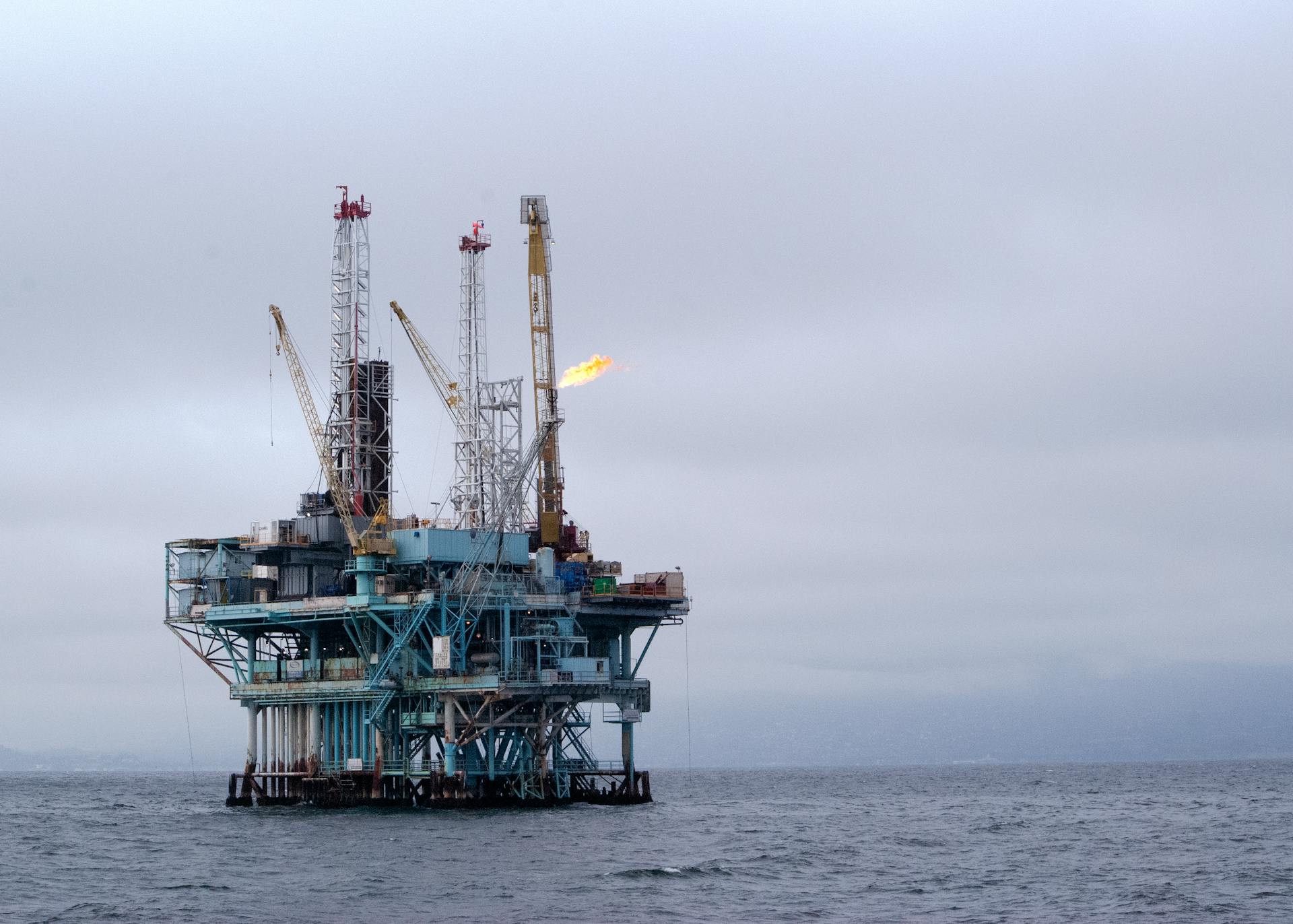 A view of an offshore oil rig in the open sea with cranes and a flare against a cloudy sky.