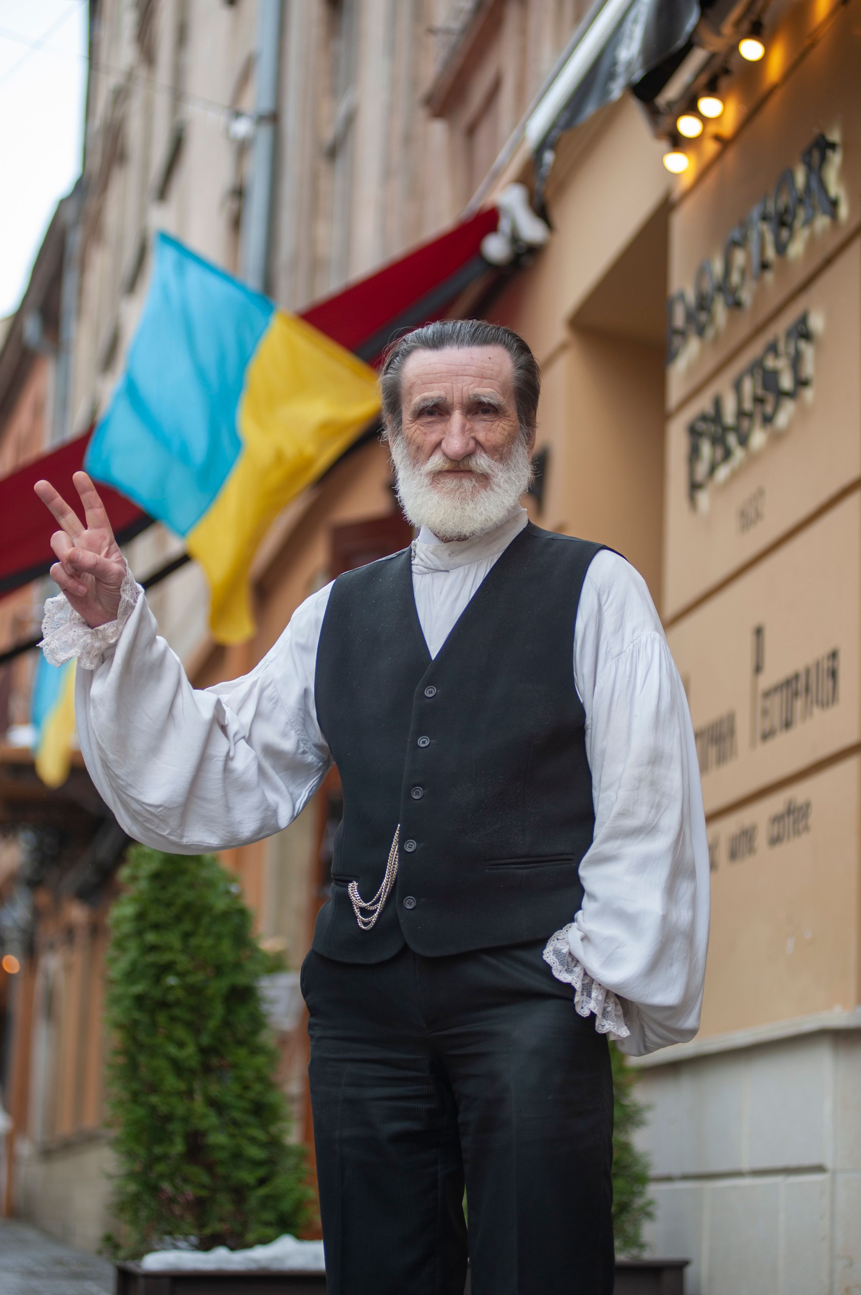 Senior man with beard and vest gestures peace sign on Lviv street by Ukrainian flag.