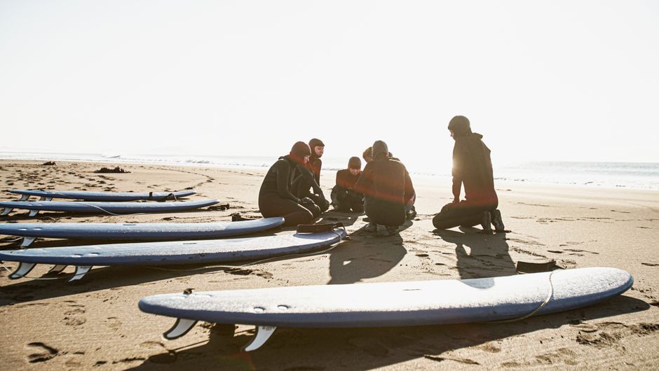 A group of surfers in wetsuits ready their boards on a sunny Kamchatka beach.